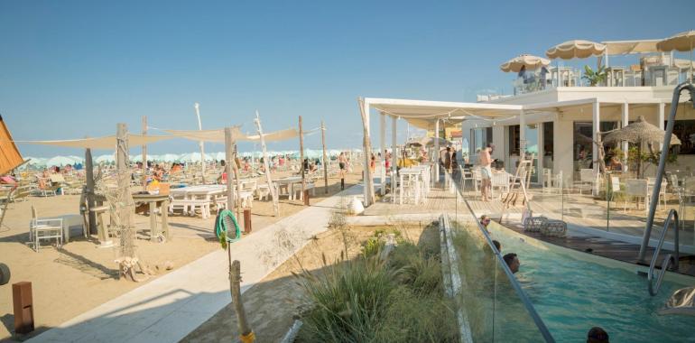 Spiaggia affollata con bar e piscina, sotto un cielo sereno.